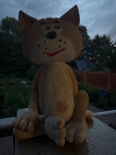 A ceramic cat figurine is seated on a stone ledge. The figurine is light brown with a cream-colored belly and paws. It has green eyes, a black nose, and a red mouth with black dots on its cheeks. The figurine is positioned outdoors, with greenery and trees visible in the background, along with a wooden fence and some structures. The sky is overcast and dark, suggesting it is either dusk or dawn. The overall scene appears to be a residential backyard or garden area, likely in the city of Kamenka, Russia, given the provided context.