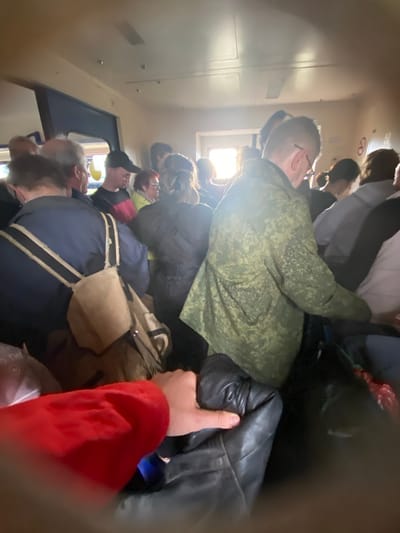 The image appears to show a crowded passenger train or bus interior. Several people are standing close together. One person is wearing a green camouflage jacket. Another person is wearing a blue jacket with white stripes. A person wearing a red garment is partially visible in the foreground. Light enters through a window on the left. The location is Vyritsa, Russia.