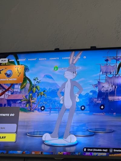 The image shows a television screen displaying the main menu of the video game Fortnite. The character Bugs Bunny is centrally positioned on the screen. Textual elements include options such as "Play," "Shop," "Locker," "Passes," "Quests," "Compete," and "Career." Other game elements include a character name and level indicator. The background depicts a tropical and pirate-themed landscape. Additional icons on the right include Chat, Emotes, and an Epic Games icon. The make and model of the screen (or TV) cannot be determined with the available information. The image doesn't pertain to Glojene, Bulgaria, and doesn't contain any people or actions within a geographic location.