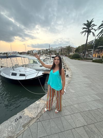 A woman stands on a paved walkway next to a marina in Port d'Alcúdia, Spain. She is wearing a turquoise tank top and brightly colored striped pants, and has her right hand raised. She is facing the camera and smiling. Several boats are docked in the water to her left, and a cityscape with buildings and palm trees is visible in the background. The sky is overcast with some clouds.