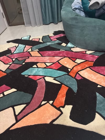 A close-up, slightly angled shot shows a section of a colorful rug with a geometric pattern. The rug features an array of colors including pink, orange, teal, and beige, separated by black outlines. A portion of a green, corduroy-textured cushion or chair back is visible in the upper left corner, with what appears to be clothing items resting on it. A window with drawn blinds is also partially visible on the left side of the frame, near the bottom. The location in the city of Arinj, Armenia is not discernable from this image.