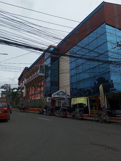 The image depicts an urban street scene in the city of Kodarapalli, India, under an overcast sky. A multi-story commercial building dominates the right and central background, featuring a modern architectural design with large sections of reflective blue glass facades and reddish-brown textured panels. The left portion of this building incorporates red horizontal cladding and an intricate red metal lattice structure visible beneath a white overhang.
At street level, the building houses an establishment with a blue sign reading "es.teh Indonesia" above its entrance. To the right of this, another shopfront displays a large yellow banner with red and green text, partially obscured, and several vertical yellow flag-like banners.
In the foreground, an asphalt road runs from the lower left towards the mid-right. A red minivan or car is partially visible parked on the far left. Along the curb, a blue scooter and multiple cycle rickshaws are parked. Further down the street, a horse-drawn carriage is stationary, with a horse and a visible driver. Numerous black electrical and communication cables crisscross horizontally across the upper portion of the frame, obscuring parts of the building and the sky. The sidewalk area features a low stone wall and some vegetation. No explicit human interactions are discernible; all vehicles appear to be parked or awaiting passengers.