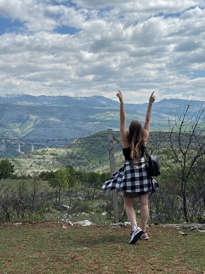 A photograph taken in the city of Gornje Mrke, Montenegro, shows a woman standing on a grassy hillside with her arms outstretched, overlooking a mountainous landscape. The sky above is filled with clouds. The woman is wearing a black top and a black-and-white checkered dress, along with sneakers. She has a small black backpack on her shoulder. A chain-link fence runs along the edge of the hillside.