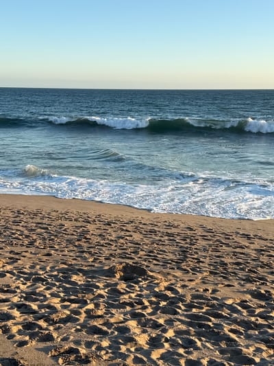 An ocean view of the Pacific Ocean with waves crashing on the shore and sandy beach in Malibu, California, United States of America. The sky is clear and blue, with a hint of yellow near the horizon. The ocean is a deep blue, with white foam from the waves. The beach is covered in sand and footprints.