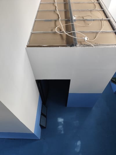 The image displays an interior view of a building located in Gadog, Indonesia, captured from an elevated perspective looking downwards. The upper portion reveals an incomplete suspended ceiling structure, consisting of a silver-colored metal T-bar grid system. Several unpainted, brownish fiberboard or gypsum panels are partially installed within this grid. White electrical wiring is loosely draped and coiled across the grid, connecting to at least two visible circular white junction boxes. Below the ceiling structure, a substantial white-painted soffit or structural beam is present. On the left, a wall section painted white angles downwards, meeting a lower wall segment painted light blue. A dark, black-framed rectangular opening, indicative of a doorway, leads into a shadowed interior space on the mid-left. Below this opening, two dark-framed rectangular window panels are visible. The floor, occupying the lower half of the image, is a uniform light blue surface, displaying multiple distorted reflections of an unseen light source. No individuals or discernible actions are present. The environment suggests an area under construction, undergoing renovation, or designed with a utilitarian aesthetic.