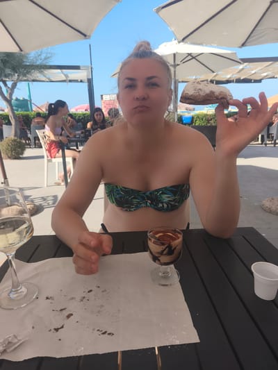 A medium shot shows a woman sitting at a dark wooden table outdoors, under a white umbrella, at what appears to be a beachside cafe or restaurant in San Salvo, Italy. She is wearing a green and black leaf-patterned bikini top and has her hair pulled up in a bun. She is holding a piece of pastry dusted with powdered sugar in her right hand, near her mouth, and has a surprised or playful expression. In front of her on the table is a glass of white wine and a dessert in a stemmed glass with chocolate drizzled on top. A crumpled piece of white paper with crumbs is also on the table. In the background, other people are visible sitting at tables and enjoying the sunny day. The environment suggests a relaxed, vacation-like atmosphere with vegetation and beach umbrellas.