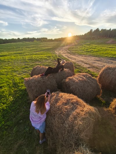 This outdoor photograph captures two women amidst a rural landscape during sunset in what appears to be Стеклянный, Russia. One woman with blonde hair, dressed in dark clothing, is relaxed and posing, lying on a cluster of large, round hay bales. Her arm supports her head as she looks towards the horizon. The second woman, seen from a high angle and from behind, has long brown hair and is wearing a light purple jacket over a striped top and shorts. She is actively taking a photo of the first woman with a white smartphone.

The scene is set in a wide, lush green field with a winding dirt path. A dense forest line stretches across the distant horizon. The sky is a mix of pale blue and white, adorned with wispy clouds. The most striking notable detail is the golden hour light of a setting sun, which is prominently visible on the horizon, casting a warm glow and long shadows across the field and hay bales. A bright lens flare emanates from the sun. A small white car is visible in the distance on the dirt path. The overall impression is one of leisure and enjoyment of the natural environment during a beautiful evening. No discernible text is visible in the image.