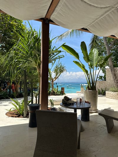 A serene view from a shaded outdoor lounge showcases a turquoise ocean dotted with boats under a clear blue sky. Lush tropical plants frame the vista. A modern gray stone table with glasses and personal items sits nearby. Sleek gray chairs surround the table, emphasizing the contemporary design. The scene evokes relaxation and coastal luxury.