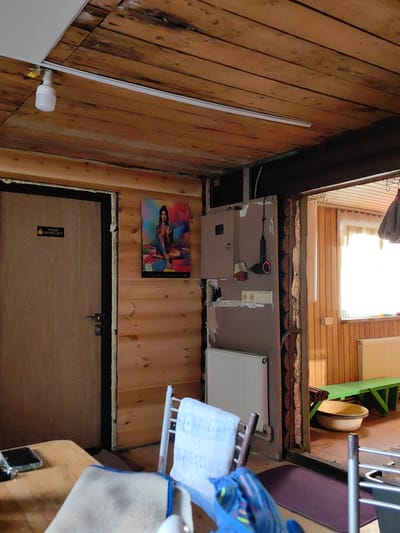 The image depicts the interior of a room with wooden walls and ceiling, possibly a sauna or bathhouse, located in the Фаніпальскі сельскі Савет, Belarus. A closed wooden door with a metal handle is visible on the left, featuring a sign with text in Cyrillic that appears to be "УХОДЯ ГАСИТЕ СВЕТ!" (Turn off the lights when leaving!). To the right of the door, a colorful poster of a woman is displayed on the wooden wall. In the center of the room, a white radiator is mounted beneath a light-colored panel. Further to the right, an open doorway leads to another room with light-colored wooden walls and a window with light-colored curtains. In this adjacent room, a white radiator is visible, along with a green wooden bench and a large, light-colored basin on the floor. Various pipes and electrical conduits are exposed on the wall. A fluorescent light fixture is attached to the ceiling. In the foreground on the left, a portion of a wooden table is visible, with a dark rectangular object, possibly a phone, resting on it. Another object, likely a chair with a white towel draped over it, is positioned in the center of the frame. Additional chairs and a dark purple mat are also present. The overall impression is one of a rustic and functional space.
