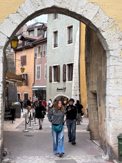 The image depicts a street scene in Annecy, France, viewed through a stone archway. In the foreground, a woman with long, wavy brown hair is walking away from the camera, wearing a striped long-sleeved shirt, denim jeans, and a turquoise crossbody bag. To her left, a young person with short, dark hair is walking towards the camera, wearing a black track jacket with white stripes, denim jeans, and holding a smartphone. Further back, several other individuals are walking and standing in the street, with one person wearing a red jacket and another in a black jacket and skirt.

The street is paved with cobblestones and lined with buildings of various colors, including peach, pink, and light gray. Some buildings have traditional wooden shutters. A yellow street lamp hangs from an ornate bracket on one of the buildings. A black sign with white lettering, partially visible, reads "Glacier Alpes" and "Depuis 1968." Another sign above a restaurant reads "Le Beignet ALPIN RESTAUR" and "Ambre" with a heart symbol. A green trash bin is visible on the right side of the image. The overall atmosphere is that of a charming European town street during the day.