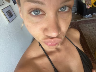 A close-up, front-facing selfie shows a woman with striking blue eyes and fair skin. Her blonde hair is pulled back, with a few strands framing her face. She is wearing a black bikini top with thin straps. Her lips are pursed, and she has a playful or coy expression. The background is slightly out of focus, but suggests an indoor setting with a textured white wall to the left. Further to the right, a dark leather armchair, a patterned rug, and what appears to be a chest of drawers are visible. The lighting suggests it is daytime, with natural light illuminating the scene. There are framed pictures on the wall behind her to the left, one of which shows two smiling individuals. The overall mood is casual and intimate.