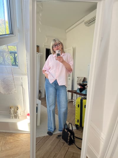 A middle-aged woman with blonde hair and glasses is taking a selfie in a mirror. She is wearing a pink and white striped button-down shirt and blue jeans. Behind her is a room with hardwood floors, a yellow suitcase, and a small black bag. A window with a printed shade is visible at the bottom of the frame. The reflection suggests the room is likely in a residential setting. The photograph offers no indication of Montélimar, France.