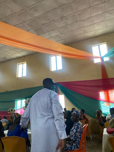 The image depicts an indoor gathering, likely a ceremony or event, in Kuru, Nigeria. The room is decorated with draped fabric in shades of orange, pink, and green hanging from the ceiling. Several windows are visible along the walls, letting in natural light. A man in a white garment is seen from behind, facing away from the camera, and appears to be addressing or interacting with a group of people seated in chairs. The seated attendees are predominantly women, many wearing vibrant headwraps and traditional clothing. They are seated at tables, some of which are covered with tablecloths. The overall atmosphere suggests a community event.