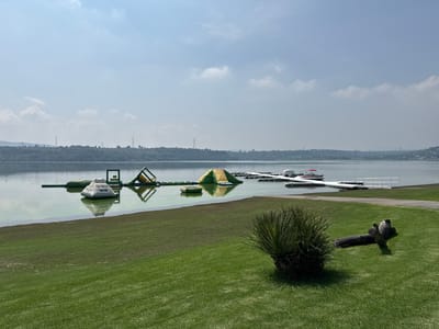 The image shows a lake with floating inflatables and a dock. A grassy hill slopes down to the water. In the foreground, a large piece of driftwood rests on the grass. In the distance, hills and power lines can be seen under a hazy sky. The location is likely a recreational area, possibly a park or resort, on the outskirts of Tepeji de Ocampo, Mexico, given the presence of floating water park structures and a dock for boats. The overall atmosphere is serene and offers a view of natural landscape combined with recreational facilities.