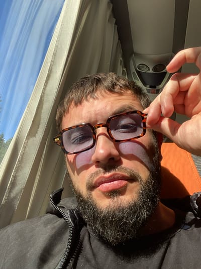 A close-up, vertically oriented image shows a man's face as he holds a pair of sunglasses in front of his eyes. The man has dark hair, a beard, and appears to be wearing a black jacket. Sunlight streams in from the right, casting shadows and highlighting the man's face and the sunglasses. The lenses of the sunglasses reflect the man's eyes and eyebrows.
In the background, to the left, a white object with two circular openings and a grille is visible, possibly a speaker or vent. The setting appears to be indoors, near a window that looks out onto a bright blue sky with wispy white clouds and green trees. The blinds or curtains are drawn, creating a striped pattern of light and shadow across the lower portion of the image. The overall lighting suggests it is daytime. The man's expression is neutral, and the focus is on the interaction with the sunglasses.