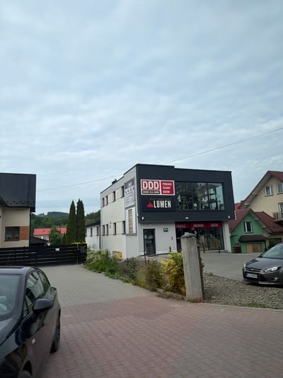 The image shows a commercial building with "DDD PODŁOGI ŚCIANY DRZWI" and "LUMEN" signage. The building is modern with a dark facade on the upper level and white on the lower level. Adjacent to it is a residential house with a red roof and a green building with a wooden garage door. A dark-colored car is parked on the right side of the image, with a license plate reading "KL1 76476". The foreground shows a brick-paved driveway and sidewalk. The background features lush green trees and hills. The scene is set in a semi-urban environment under a cloudy sky.