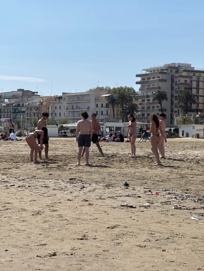 This photograph captures a beach scene in Anzio, Italy. The foreground features a sandy beach with visible litter. Groups of people populate the beach, some walking and others relaxing. In the middle ground, several buildings and palm trees are visible, indicating the proximity of the town to the beach. The sky is clear and blue, suggesting a sunny day.