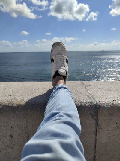 The image shows a person's right leg resting on a concrete wall overlooking the ocean. The person is wearing light blue jeans and a white sneaker with black detailing. The leg is positioned so that the foot is pointing upwards, with the toes facing the viewer. The ocean is visible in the background, with a bright blue sky and white clouds. The location is the coastal city of Parede, Portugal, at coordinates 38.685 latitude and -9.353 longitude. The scene is peaceful and serene, with a sense of calm and relaxation. The person is likely enjoying the view and the fresh air. The image has a casual and spontaneous feel.