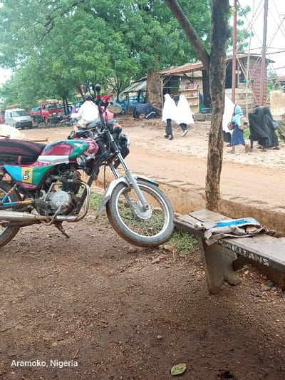 A red motorcycle with a green, white, and blue stripe is parked on a dirt road near a wooden bench with the inscription "ISANILAYS". Several people are walking in the distance, including women wearing long, white robes. The image was taken in Ilobu, Nigeria, located at 7.835 latitude and 4.479 longitude. A tree trunk and a large branch extend from the left foreground of the image into the middle ground. The road and the ground around the motorcycle and bench are covered in dirt and small rocks. The sky is overcast and there are several trees in the background. The overall scene is a typical street scene in a rural area of Nigeria.
