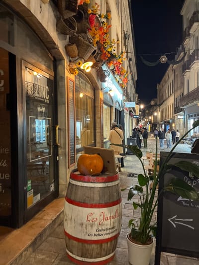 A nighttime street scene shows a European-style street lined with buildings decorated for autumn. A barrel is decorated with the words "Les Jamelly" and holds a pumpkin in a cardboard box. A chalkboard with an arrow pointing down is next to the barrel. "LE PETIT ZINC" is displayed above a window. People stroll in the distance, under soft lighting. Decorative lights and floral arrangements adorn the building facades, and potted plants add to the charm.