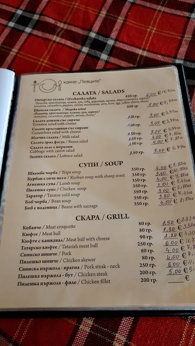 The image shows a menu from a restaurant named "Ханче „Певците“" (Hanched "Pevtsite"), likely located in Pevtsite, Bulgaria. The menu is presented in Bulgarian and English, divided into sections for Salads, Soups, and Grill. The items listed include various salads like Ovcharska and Shopska, soups such as Tripe and Lamb soup, and grill items like meat croquettes and chicken skewers. Prices are listed in Bulgarian Lev (лв) and Euros (€). The menu is open on a table with a red and black plaid tablecloth visible in the background, suggesting an indoor setting.