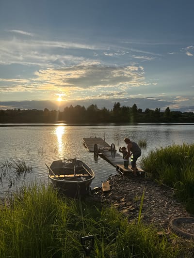 This image depicts a sunset scene at a lake, possibly located near Sitno, Russia. In the foreground, a dark-colored boat is tied to the grassy shore. A wooden dock extends into the water, with two figures, an adult and a child, standing on it. The adult, wearing a striped shirt, appears to be interacting with the child. The lake reflects the warm glow of the setting sun, which is visible in the distance behind a line of trees. The sky is filled with scattered clouds, illuminated by the sun. The overall atmosphere is serene and peaceful, characteristic of a rural or natural setting.