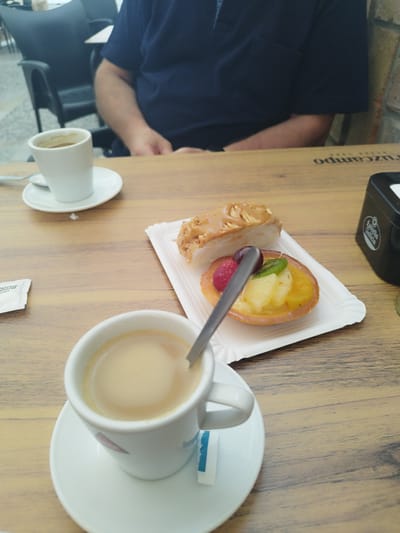 In a cafe in Rota, Spain, a close-up, eye-level shot captures a wooden table with two cups of coffee and two pastries. In the foreground, a white ceramic coffee cup sits on a matching saucer, filled with light brown coffee and topped with a frothy layer. A spoon rests inside the cup. To the right of the cup, a white paper napkin with blue accents is partially visible.

In the mid-ground, a white paper plate holds two pastries. One is a slice of cake with a glazed topping and thinly sliced almonds. Next to it, a small tart is filled with colorful fruit, including raspberries, kiwi, pineapple, and possibly orange segments, with a grape on top. A silver spoon is placed across the tart.

In the background, another white ceramic coffee cup rests on its saucer, also filled with coffee. A person, only their torso and arms are visible, wearing a dark blue shirt, is seated at the table. They are partially obscured by the coffee cup in the foreground. The cafe environment is suggested by the presence of other seating and furniture, possibly a brick wall in the far background. The overall lighting suggests it is daytime.