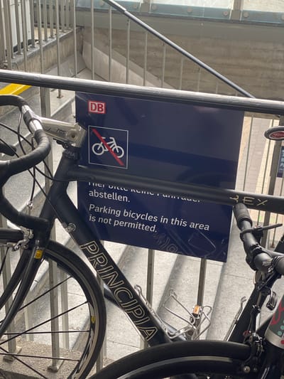 A bicycle is parked next to a Deutsche Bahn (DB) sign that reads "Parking bicycles in this area is not permitted," likely on a staircase in a public transportation area in Heidelberg, Germany. The bicycle, with "PRINCIPIA" on its frame and "REX" visible on the stem, appears to be a road bike with a Mavic wheel. The sign is mounted on a metal pole, presumably part of the staircase railing. The background shows a stairway and possibly a glass or metal structure above or beside the staircase. The angle and orientation of the image suggest it was taken from a slightly elevated position relative to the sign and the bicycle.
