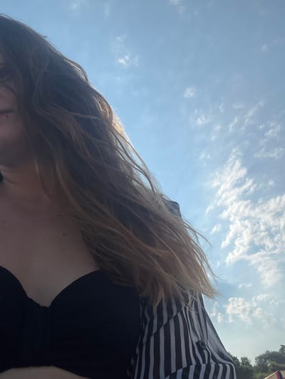 The media file depicts a person's upper body and head from a low-angle perspective, with the sky and clouds dominating the background. The person has long, light brown hair that is blowing in the wind, suggesting an outdoor setting with a breeze. They are wearing a black top with a striped pattern visible on the shoulder area. The sky is a bright blue with scattered white clouds, indicating it is daytime, likely sunny with some cloud cover. The overall impression is casual and relaxed, possibly taken on a warm day. The scene is set outdoors, with the sky and clouds being the most prominent elements. There are no visible objects or text in the frame that provide further specific context about the location or activity.