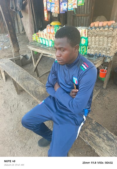 A man in a blue tracksuit with an Italian flag patch sits on a weathered wooden bench. He is next to a makeshift stall with food items, including eggs, stacked high. The stall has corrugated metal walls and a variety of snacks hanging. The area appears to be outdoors, with a dirt ground and various objects scattered around. He has his arms folded and a thoughtful expression.