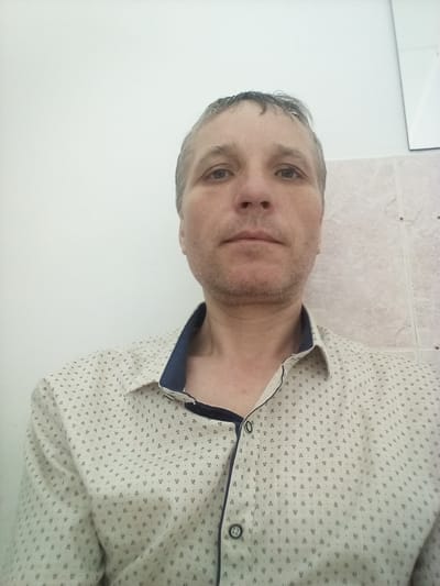 The media file is a selfie of a middle-aged man with short, graying hair and light stubble. He is wearing a light-colored collared shirt with a subtle, dark pattern of small, abstract shapes. The shirt has a dark blue trim around the collar and placket, and two dark buttons are visible. He is looking directly at the camera with a neutral expression.
The background appears to be a room with plain, white walls and a section of pale pink tiled wall to the right. The lighting is bright, suggesting it is daytime, but no specific weather conditions are discernible. The overall setting seems to be a domestic or indoor environment, possibly a bathroom or hallway, given the tiling. There are no other people or objects of note in the frame. No text is visible in the image. The overall impression is a candid, informal portrait.