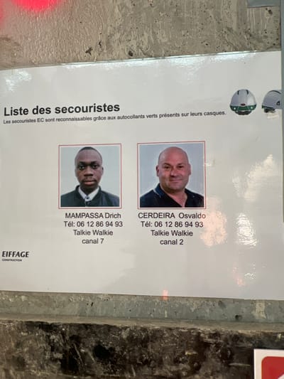A laminated white sign, titled "Liste des secouristes" (List of first responders), is affixed to a coarse, gray concrete surface in Aulnay-sous-Bois, France. The sign displays two passport-style headshots: on the left, MAMPASSA Drich, a male with dark complexion, wearing a black jacket over a white shirt; on the right, CERDEIRA Osvaldo, a bald male with light complexion, wearing a dark polo shirt. Below each photo, their names are listed, followed by a shared telephone number "06 12 86 94 93", and individual "Talkie Walkie" channels (7 for MAMPASSA, 2 for CERDEIRA). An explanatory text above the photos reads, "Les secouristes EC sont reconnaissables grâce aux autocollants verts présents sur leurs casques" (EC first responders are recognizable by the green stickers on their helmets), accompanied by two helmet illustrations, one showing green markings. The "EIFFAGE CONSTRUCTION" logo is located in the lower-left corner of the sign. A faint red mark is visible on the concrete surface above the sign's top left edge.