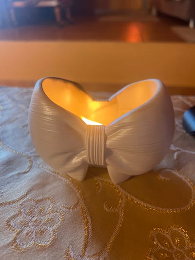 The scene depicts an indoor environment within the Gerakas Municipal Unit, Greece, under low ambient light conditions. A white, bow-shaped decorative object is centrally positioned on a light gold fabric surface. The object has a matte, slightly textured finish with visible horizontal striations across its surface, consistent with a molded or 3D-printed item. A warm, diffused orange-yellow light emanates from within the object's hollow interior. The fabric beneath the object is adorned with an intricate, repeating pattern of gold-colored embroidery. To the right, a dark, out-of-focus object is partially visible in the mid-ground. The background is blurred, showing warm, muted orange-brown tones indicative of an interior wall or furniture. No individuals are present, and no discernible actions or interactions are depicted.