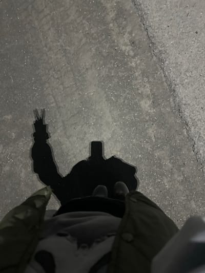 The image is a top-down shot of a shadow on a concrete surface, likely a sidewalk or road, in Pionersky, Russia. The shadow depicts a person holding a smartphone and making a peace sign with their hand. The person is wearing a jacket and appears to be standing. The concrete is textured with small pebbles and a crack runs diagonally across the frame. The lighting suggests it is evening or night.