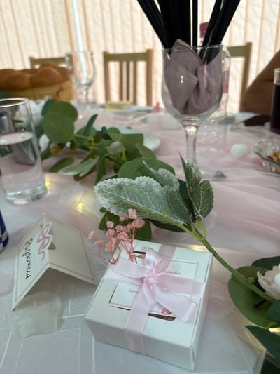 This image captures a close-up, eye-level view of a table setting arranged for an event in Strazhitsa, Bulgaria. The table is covered with a white tablecloth overlaid with sheer pink fabric, underneath which small, warm-toned fairy lights are visible.

In the foreground, a white rectangular favor box, approximately 10x8 cm, is tied with a pink ribbon and bow, resting on the pink fabric. A white label on the box is partially obscured by the ribbon, with visible Cyrillic text including "ски бал". To its left, a white tent-fold place card, approximately 8 cm tall, features pink design elements and vertically oriented Cyrillic text that appears to read "Рожден ден" (Birthday). Artificial floral decorations, comprising green leaves and clusters of small pink flowers resembling baby's breath, are placed around these items.

Further back on the left, a clear glass tumbler filled with water stands on the table. A small blue and white patterned object, possibly a miniature bottle or vase, is partially visible on the far left edge. In the midground, artificial green foliage resembling eucalyptus branches extends across the table. Towards the upper right, a clear stemmed glass, approximately 15 cm tall, contains a folded light pink or purple napkin and several black drinking straws. Behind this glass, a clear plastic bottle with a pink cap is visible. On the far right, a small fluted glass bowl holds a quantity of nuts or similar snacks.

The background reveals light-colored vertical window blinds or curtains. Two wooden chairs with slatted backs are partially visible, indicating seating arrangements. A blurred, braided loaf of bread is situated on a wooden stand in the upper left background. A human arm or shoulder is faintly visible on the far right edge of the frame. No specific actions or interactions are depicted beyond the arranged setting.