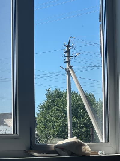 The image, taken from inside a building through a window, offers a view of an outdoor scene. The scene is dominated by a bright blue sky and an electrical pole with several wires extending from it. The pole itself appears to be made of concrete and is tilted at an angle. Behind the pole, a dense green tree provides a backdrop. In the foreground, on the windowsill, is a folded piece of light-colored fabric, possibly a towel or blanket, with a textured surface. To the left of the window frame, part of another building with a brick facade and a fence is visible. The scene is consistent with an urban or semi-urban environment, and the presence of electrical infrastructure and buildings suggests it is located within a populated area. This image is relevant to the city of Zudilovo, Russia.