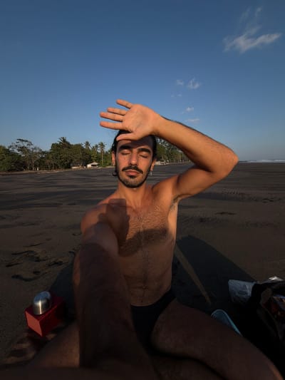 This image captures a man on a dark sand beach in Selemadeg, Indonesia, under a clear blue sky with minimal white clouds. The man, with dark hair, a beard, and a mustache, is bare-chested and wears black swim briefs. He has a cross earring in his left ear. His eyes are closed, and his right hand is raised, covering his forehead to shield his eyes from the sunlight. His left arm is extended towards the camera, indicating a selfie. Long shadows are cast to the left of the man, suggesting the sun is low on the right side of the frame. In the background, a dense line of green tropical trees, including palm trees, runs along the beach, with small, simple structures visible amongst them. Further to the right, the ocean is visible with small waves. On the sand to the man's left, a red rectangular object topped with a silver, domed cap is present. To his right, a white plastic bag and other personal belongings are partially visible.
