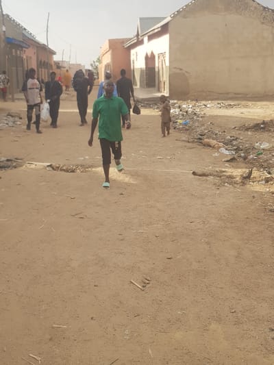 In a residential area of Dutsinma B, Nigeria, a group of people are walking along a dirt road. One person in the foreground is wearing a green shirt and dark shorts. Several other people are walking behind them, dressed in casual clothing. The road is unpaved, and there is some trash and debris along the sides. The buildings in the background appear to be simple, one-story structures. The sky is overcast, suggesting it might be early morning or late afternoon.