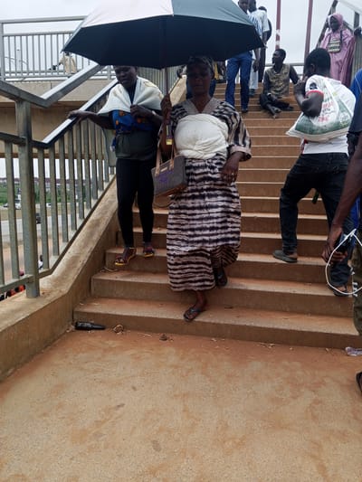 A group of people are on an outdoor pedestrian overpass in Sauka, Nigeria. The overpass has concrete stairs and metal railings. In the foreground, a woman is holding a black umbrella over herself and another person, who is wearing a white cloth around their shoulders. The woman is wearing a striped dress and dark leggings, and the other person is wearing a blue and green patterned garment. To the right, a man in a white t-shirt with writing on it is walking up the stairs, carrying a white bag. Behind him, another man in dark pants is also ascending. Further up the stairs, a man is sitting down with a broom, and a woman in a pink hijab is standing. A dark bottle and some debris are on the lower steps of the overpass. The sky is overcast.