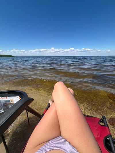 The image depicts a person sitting in a red beach chair partially submerged in shallow, clear water. The person's legs and bikini bottoms are visible. The water is calm with small ripples and reflects the bright blue sky above. In the distance, a strip of green vegetation lines the horizon, separating the water from the sky, which is dotted with scattered white clouds. The scene appears to be a peaceful lakeside or seaside setting.