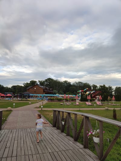 This media file captures a scene at a water park on an overcast day, likely in the late afternoon or early evening. In the foreground, a young girl with blonde hair pulled into pigtails is walking across a wooden bridge. She is wearing a striped t-shirt and shorts. The bridge has wooden railings with decorative wooden supports and hanging flower baskets.
Beyond the bridge, the water park unfolds. A pathway leads towards a large wooden building with a dark roof. To the right of the building, colorful water slides are visible, indicating a recreational setting. Numerous pink beach umbrellas are dotted across a well-maintained lawn, with lounge chairs placed beneath them, suggesting areas for relaxation. Some trees and dense foliage line the background, framing the park. The sky is filled with heavy, grey clouds, creating a subdued lighting condition. The overall atmosphere appears calm and relaxed, with no explicit indication of intense activity, though the presence of the water park and lounge areas suggests it is a place for leisure. The location is contextually identified as Shcherbani, Ukraine.