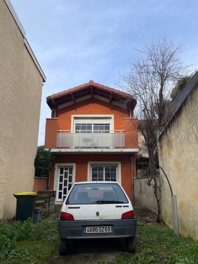 A white Peugeot 106 hatchback, displaying the French license plate "4686 SZ 69", is parked facing away from the viewer on an unpaved, grassy patch of ground in Villeurbanne, France. The vehicle's rear bumper is dark gray. Behind the car stands a two-story house with terracotta-orange stucco walls and a red-tiled gabled roof with exposed wooden eaves. The upper floor features a wide window with a partially open white rolling shutter, fronted by a balcony with frosted glass and metal railings. A small round table and two chairs are visible on the balcony. The lower floor presents a white door with multiple rectangular glass panes on the left and a large white-framed window on the right. To the immediate left of the house, a tall, plain beige wall of an adjacent building is visible, alongside a dark green refuse bin with a yellow lid. Green foliage grows against the left side of the house. To the right, a tall, weathered yellow wall borders the scene, with a bare deciduous tree situated between it and the orange house. A black pipe encased in a gray conduit runs vertically up the right wall, curving over near the top. The ground is covered with patchy green grass and bare soil. The sky is light blue with scattered white clouds. No individuals are present in the scene.