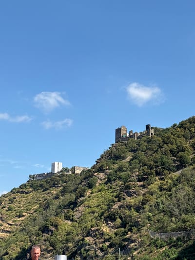 The image shows a view of Boppard, Germany, focusing on the landscape and historical structures on a hillside. The foreground is occupied by a man's head and shoulders, wearing a dark shirt. He is looking downwards and slightly to his left. Behind him, a metal cylindrical object is visible. The middle ground features a steep, vegetated hillside with a mix of trees and shrubs. Two structures are prominent on the hillside: on the left, a white modern building with multiple floors and windows, and to its right, a more ancient stone structure that appears to be a castle or fortress ruin, characterized by weathered stone walls and towers. The background is dominated by a clear blue sky with a few scattered white clouds. The overall scene suggests an outdoor location on a sunny day, likely overlooking the town of Boppard from a higher vantage point.