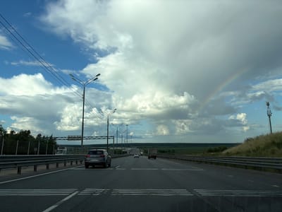 The image shows a wide, multi-lane highway with a concrete barrier on the left and a grassy embankment with a telecommunications tower on the right. Several vehicles are visible on the road, including a dark gray SUV in the foreground, a white truck, and other cars further down the highway. The sky is filled with dramatic, heavy clouds, and a faint rainbow is visible on the right side of the frame. The landscape beyond the highway appears to be agricultural fields and trees. The scene suggests a travel or transportation context, possibly on a cloudy day with recent or impending rain, given the presence of the rainbow. The location is identified as Lipitsy, Russia.