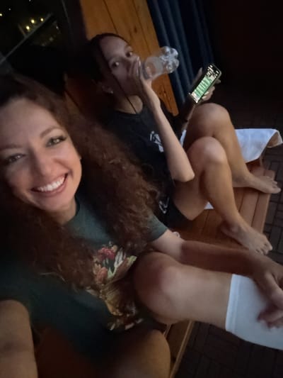 Two women are posing for a selfie in a dark environment, possibly outdoors on a wooden deck or platform. One woman, closer to the camera, is smiling with red curly hair wearing a floral-patterned top. The other woman, further away and partially obscured, is holding a water bottle and a smartphone, with her legs raised onto a wooden structure. One woman has a white towel covering her legs. The background is dark, suggesting the photo was taken at night or in a dimly lit area. It is not possible to tell that the image was taken in Bắc Hà, Vietnam from the visual information provided.