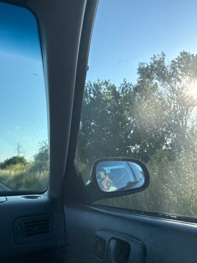 The image, captured from inside a vehicle, shows a view through the car's windshield and side mirror. The windshield reveals a bright blue sky and trees in the distance, with some sunlight glinting through the leaves. A power line tower is visible on the left side of the frame. The side mirror reflects the interior of the car and a person wearing sunglasses and holding a phone, seemingly taking a selfie. The dashboard and door panel of the car are visible in the foreground. The scene appears to be a roadside environment, possibly on a highway or rural road, given the presence of trees and vegetation. The location of Cherni Vrah, Bulgaria, is provided as context, suggesting the image was taken in that area.