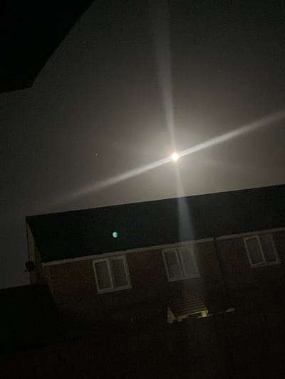 The image shows a night scene with a bright light source, possibly the moon, illuminating the sky. A building stands to the right, featuring several windows. The location is Tonbridge and Malling, United Kingdom, as the prompt specified this.