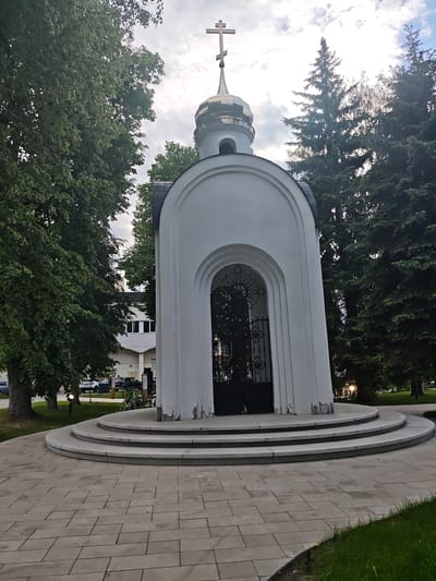 The image depicts a white, arched structure resembling a small chapel or shrine, topped with a golden dome and a Christian cross. It stands on a circular platform with three steps, and is surrounded by trees. The foreground consists of a paved walkway. In the background, a modern building and several cars are visible. This structure is located in Noginsk, Russia.