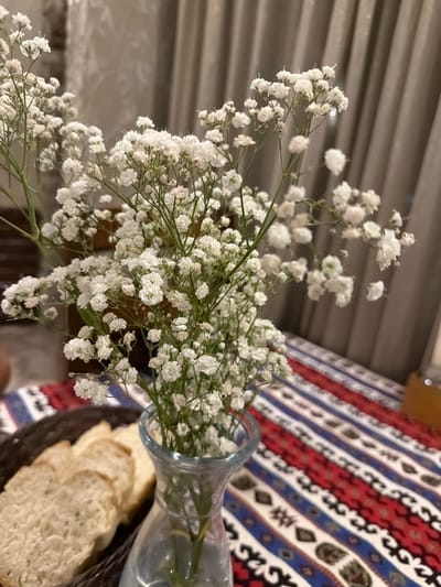 In Kumbor, Montenegro, a close-up shot captures a delicate arrangement of white baby's breath flowers in a clear glass vase. The vase sits on a table adorned with a vibrantly patterned tablecloth featuring red, blue, and white stripes with intricate ethnic motifs. To the left of the vase, a dark woven basket holds several slices of bread, suggesting a meal setting. In the background, neutral-toned curtains hang, creating a soft, diffused light that highlights the floral display and the textures of the table setting. The overall scene evokes a sense of simple elegance and domestic tranquility.