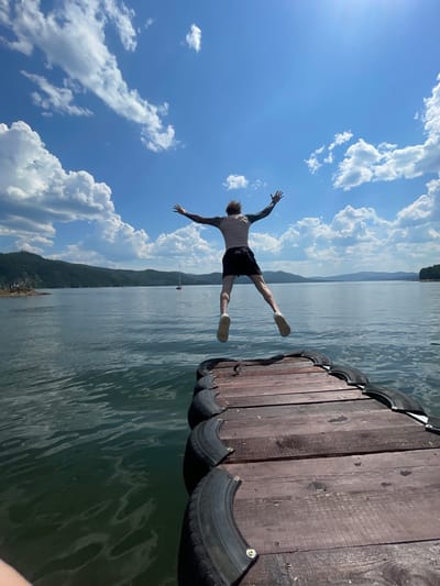 A person is captured mid-air, jumping from a wooden dock into a body of water. The image is oriented vertically, with the sky taking up a significant portion of the frame on the left. The person, seen from behind, is wearing black shorts and a light-colored t-shirt, with their arms and legs outstretched as if in a dive. They have tattoos on their left arm.
The dock, made of weathered wooden planks, occupies the right side of the image. It appears to be reinforced with sections of tires along its edge. The water is calm, reflecting the bright blue sky and fluffy white clouds. In the distance, a tree-covered hillside stretches across the horizon, with a hazy mountain range beyond. A small sailboat can be seen on the water.
The weather is sunny with scattered clouds, suggesting a clear, pleasant day. The overall mood is one of exhilaration and freedom associated with summer activities and water sports. The scene is likely set during daylight hours, given the brightness and shadows. The context provided places the location in Divnogorsk, Russia. No visible text is present in the image.